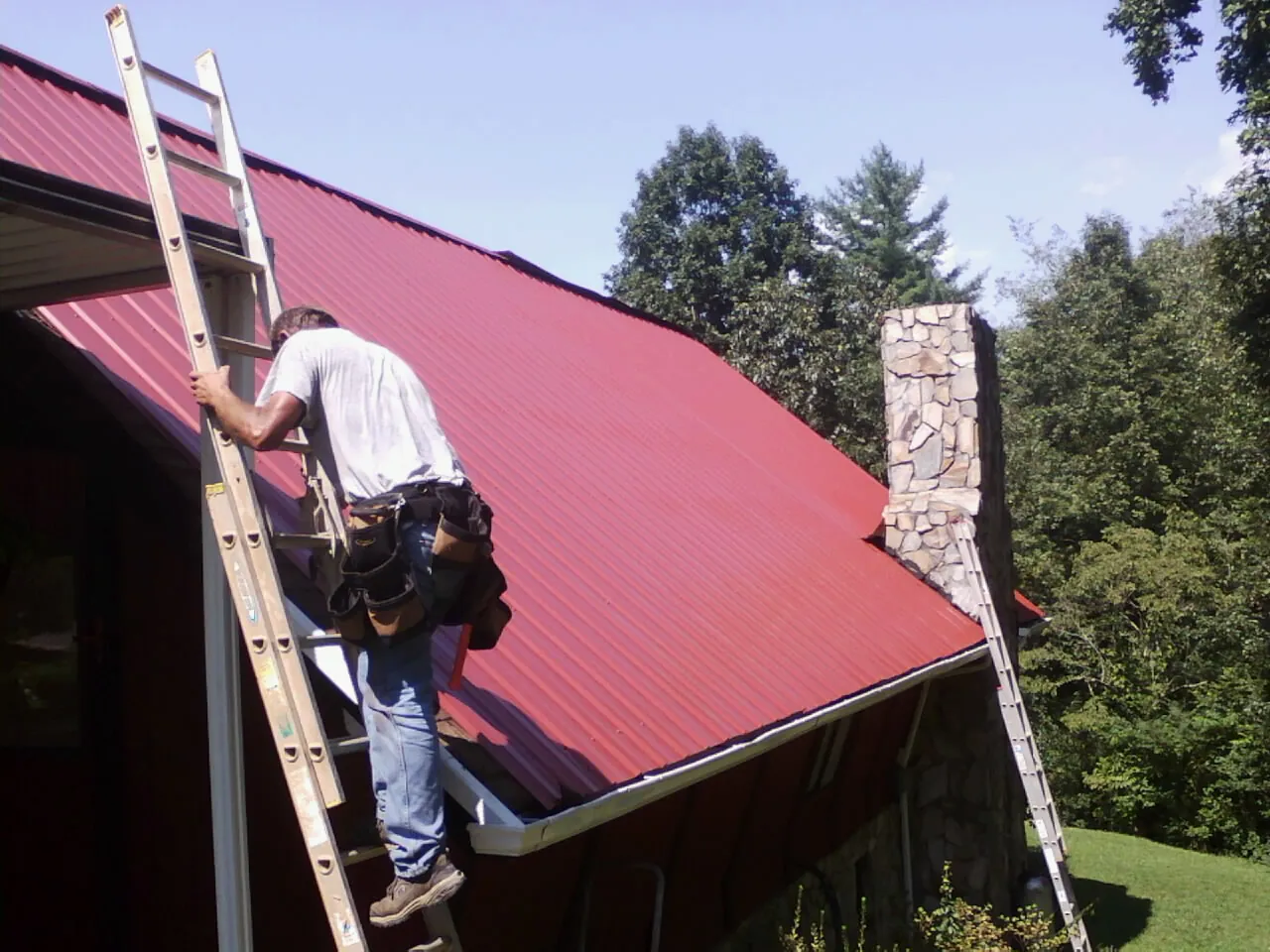 Roofer on a ladder beside a red metal roof and stone chimney — Soffit & Fascia Repair by Roofing Brothers in Newton
